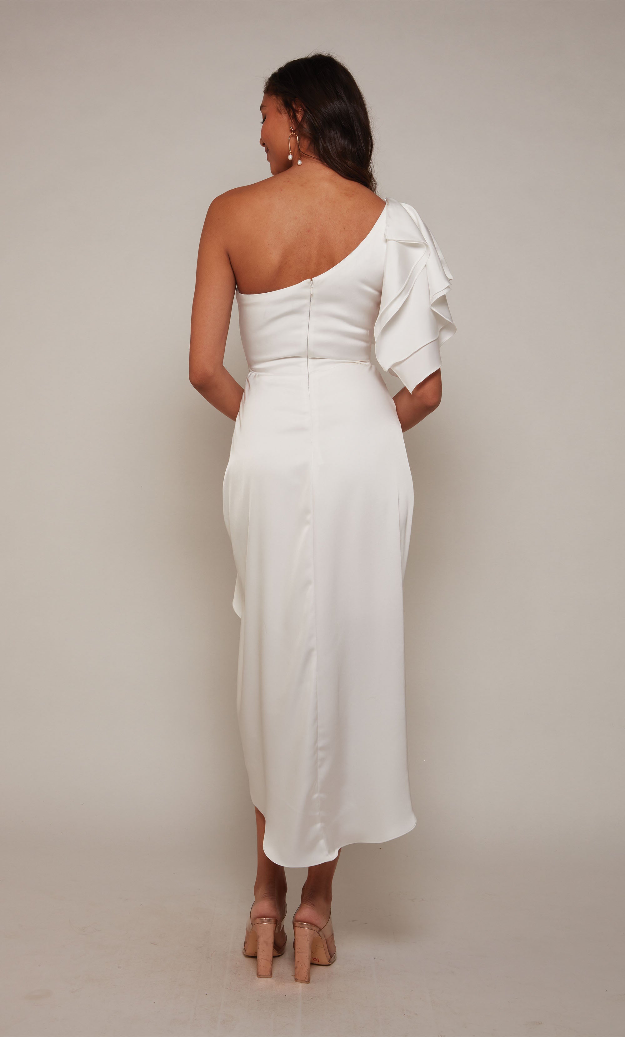 A white one shoulder cocktail dress with an asymmetrical hemline, ruffle shoulder, and cascading front drape.