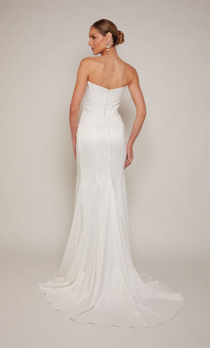 A strapless, corset wedding dress with a ruched waist, an elegant side slit, and detachable side train for a dramatic effect. The back of the dress is adorned with satin covered buttons.